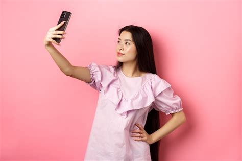 세련 된 아시아 여자 Selfie를 복용 하 고 웃 고 소셜 미디어 사진을 위해 포즈 분홍색 배경에 드레스에 서 서