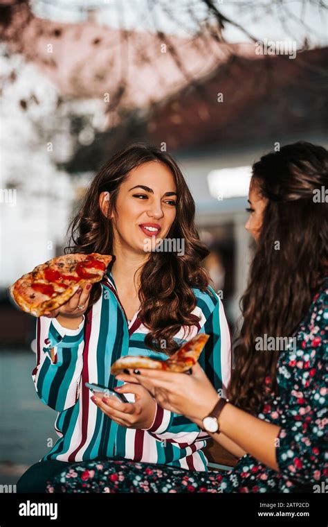 Portrait Of Two Beautiful Young Women Talking And Eating Pizza Outdoors Having Fun Together