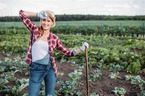 Beautiful Young Female Farmer In Hat Holding Hoe And Working On Field