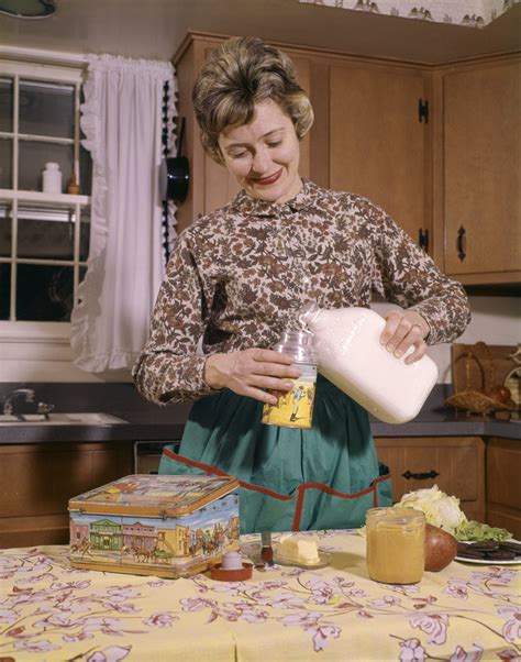 1960s Woman Housewife Mother Wearing Apron In Kitchen Pouring Milk Into