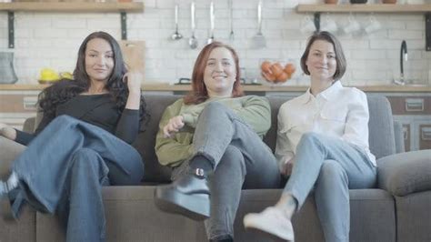 Three Cheerful Women Crossing Legs And Laughing Sitting On Couch With Kitchen At Background
