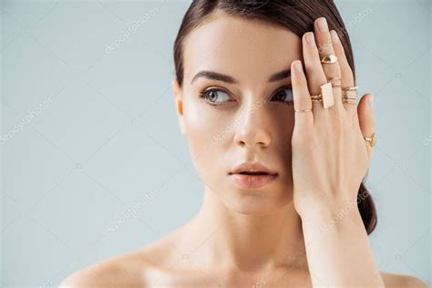 mujer desnuda joven con maquillaje brillante y anillos de oro escondiendo la cara detrás de la