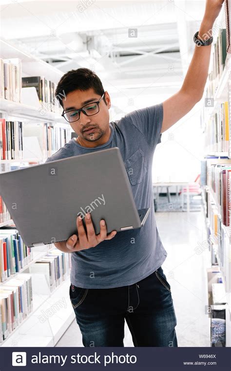 Student Holding Laptop And Reaching For Book On Shelf In Library Stock
