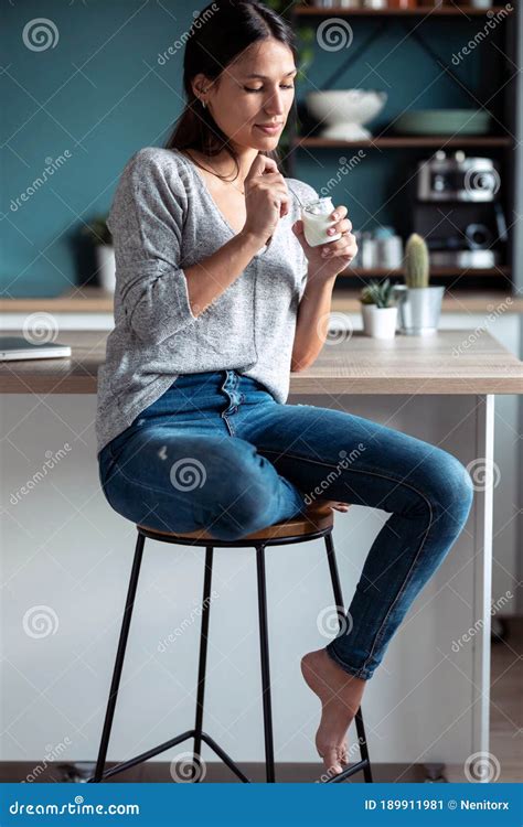Uśmiechnięta Młoda Kobieta Jedząca Jogurt Siedząca Na Stołku W Kuchni W Domu Obraz Stock Obraz
