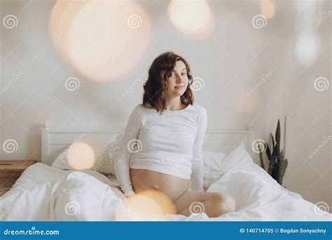 Happy Pregnant Woman In White Holding Belly Bump And Relaxing On White Bed At Home Stylish