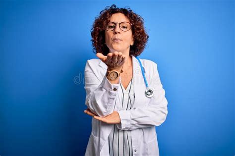 middle age curly hair doctor woman wearing coat and stethoscope over blue background looking at