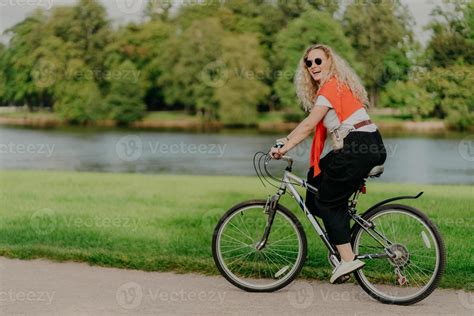Outdoor Shot Of Happy Blonde Curly Woman Wears Sunglasses Dressed In