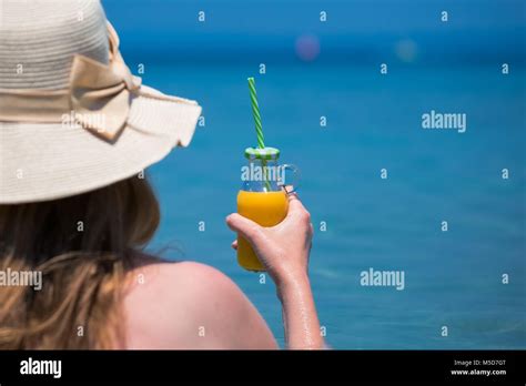 Rear View Of Female In Sunhat Holding Glass Bottle Of Orange Juice With Drinking Straw Against