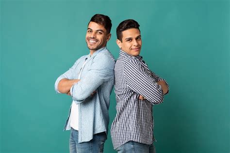 Two Arab Male Friends Standing Back To Back Over Turquoise Background