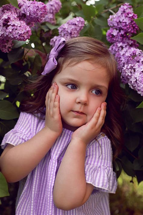 Three Year Old Girl Stands In Lilac Bushes In A Dress And A Bow