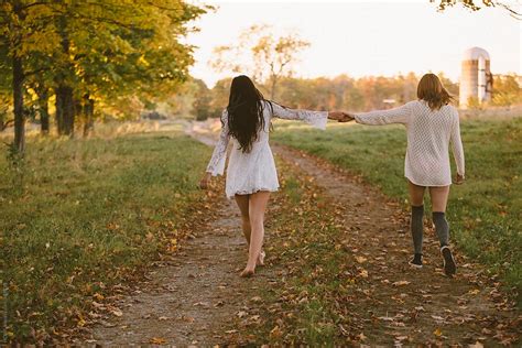 Two Young Girls Holding Hands And Walking Down A Dirt Path By Stocksy