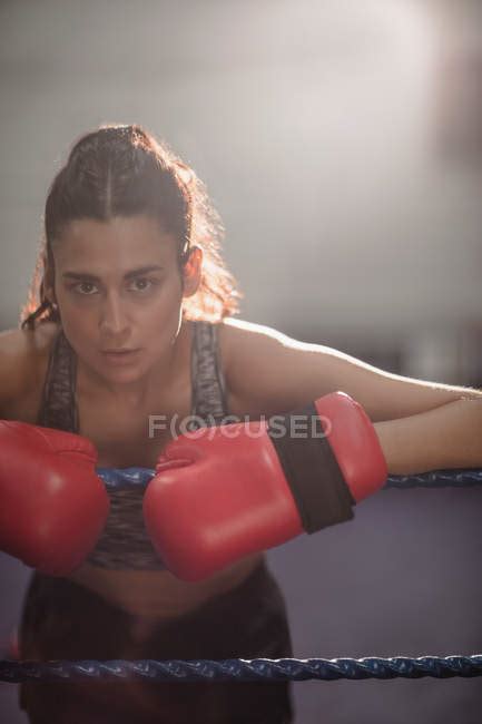 Portrait Of Female Boxer In Gloves Leaning On Boxing Ring Rope At