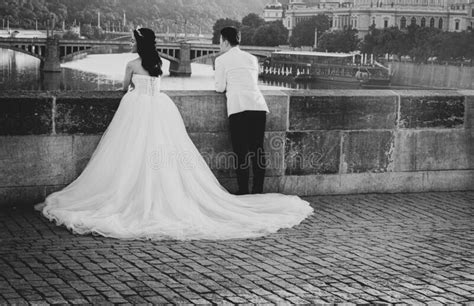 Just Married Couple In Wedding Dress And White Jacket Lean On Side Of Charles Bridge Looking