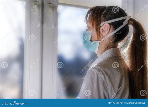 A Woman In A Medical Facial Mask Looks Out The Window While Remaining