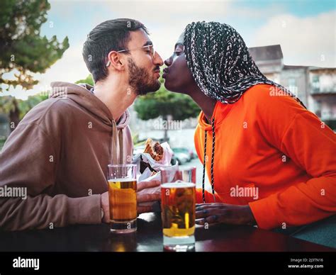 romantic biracial couple kissing sitting at the kiosk drinking beers eating sandwiches stock