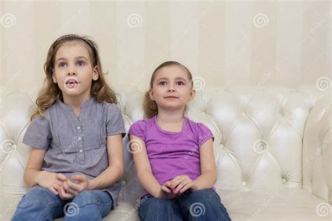 duas meninas bonitos que sentam se em um sofá e em uma tevê de observação imagem de stock