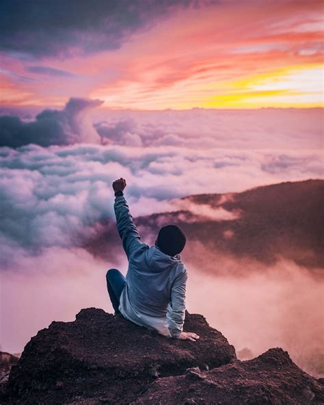 Man Sitting On Mountain Cliff Facing White Clouds Rising One Hand At