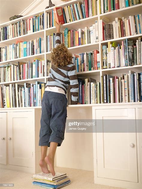 Boy Standing On Books Reaching For Top Book Shelf Rear View High Res