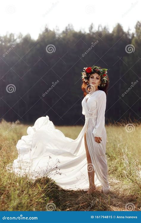 Girl In A Long Dress Stands In A Field With A Wreath On Her Head And A Bouquet Of Flowers In Her