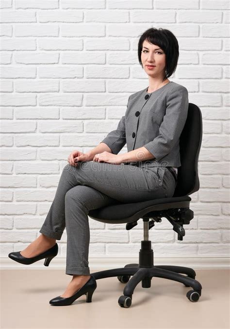 The Business Woman Sitting On A Chair Dressed In A Gray Suit Poses In Front Of A White Wall