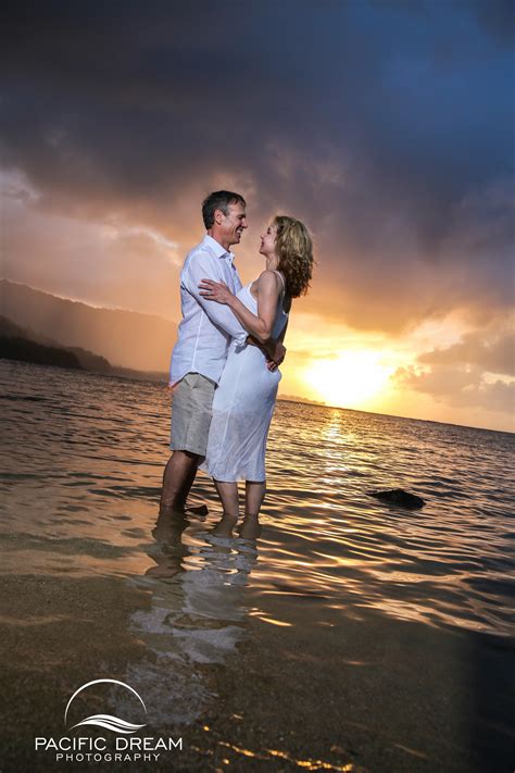 A Man And Woman Standing In The Water At Sunset With Their Arms Around