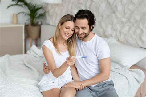 happy husband and wife rejoicing positive pregnancy test sitting on bed in bedroom interior