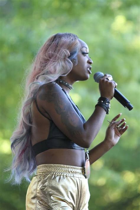 a woman with pink hair and tattoos on her chest singing into a microphone in front of trees