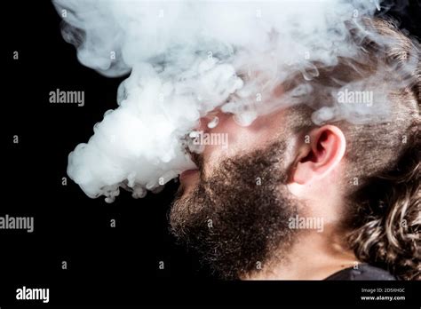 Man With A Leafy Beard Exhales Smoke Through His Mouth And Surrounds Him On Black Background