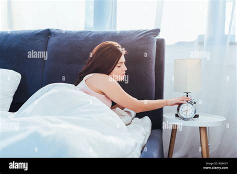 Side View Of Young Woman Turning Off Alarm Clock In Bedroom During Morning Time At Home Stock