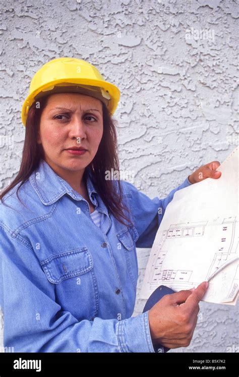 Hispanic Woman Engineer Wearing Hard Hat Examines Blueprints Of Project