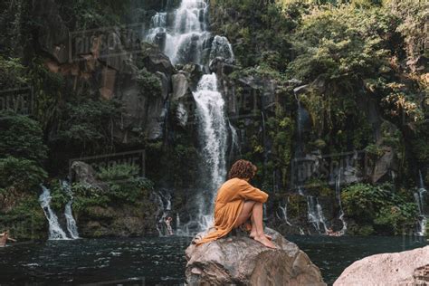 Side View Of Woman Sitting On Rock Against Waterfall In Forest Stock