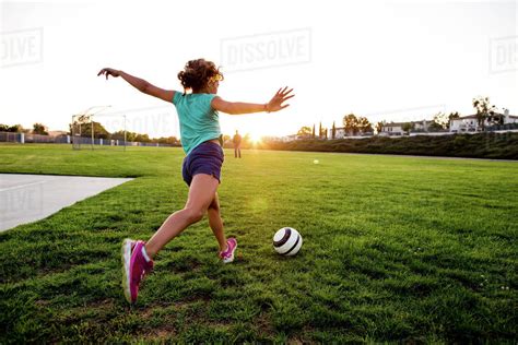 Full Length Of Girl Playing Football On Grassy Field During Sunset
