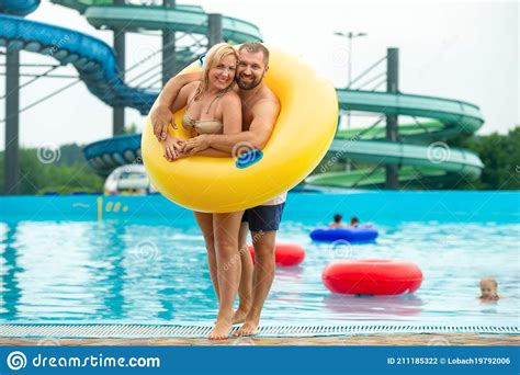 A Man And A Woman In A Rubber Inflatable Circle Stand Against The Background Of A Summer Water