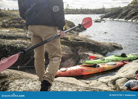 Man With A Paddle Stands On The Rocky Shore Of The Lake Against Stock