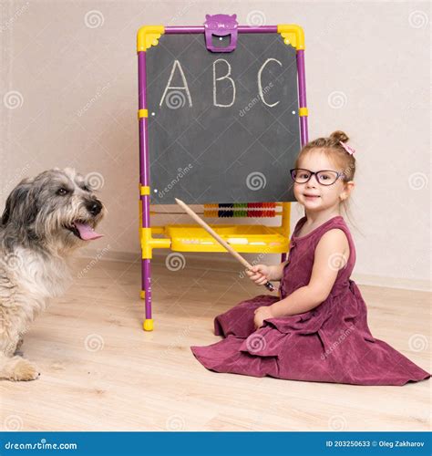 A Little Girl Teacher In A Dress And Glasses Sits On The Floor Near The