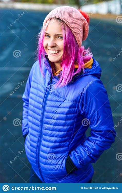 Outdoor Portrait Of Smiling Funny Woman In Blue Down Jacket And Pink Hat Posing Against Urban