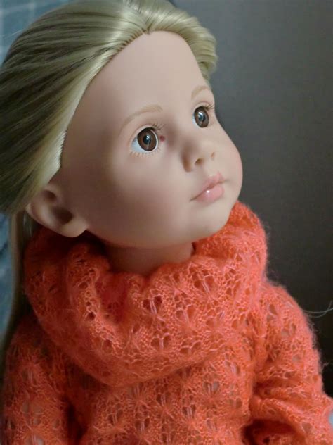 A Doll With Blonde Hair Wearing An Orange Knitted Sweater And Scarf On