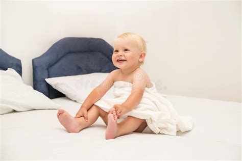 Cute Baby Smiling Under A White Towel And Sits On The Bed Stock Photo