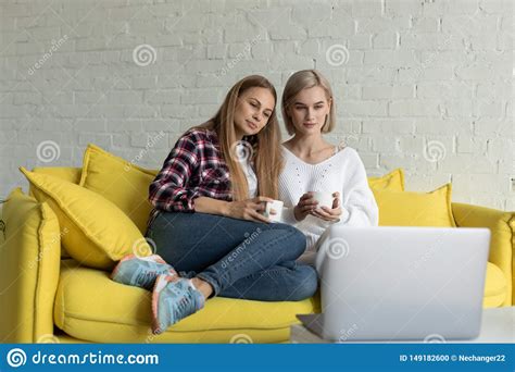 Two Beautiful Lesbian Female In Casual Clothes Sitting Together On Yellow Sofa At Home Stock