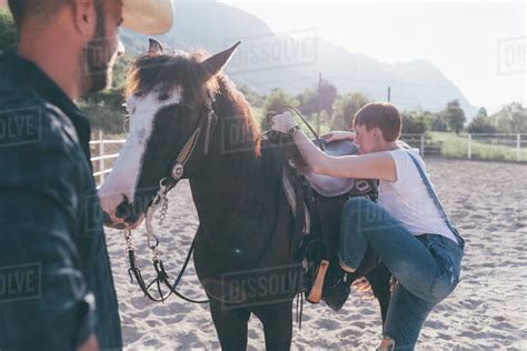 Young Woman Mounting Horse In Rural Equestrian Arena Stock Photo