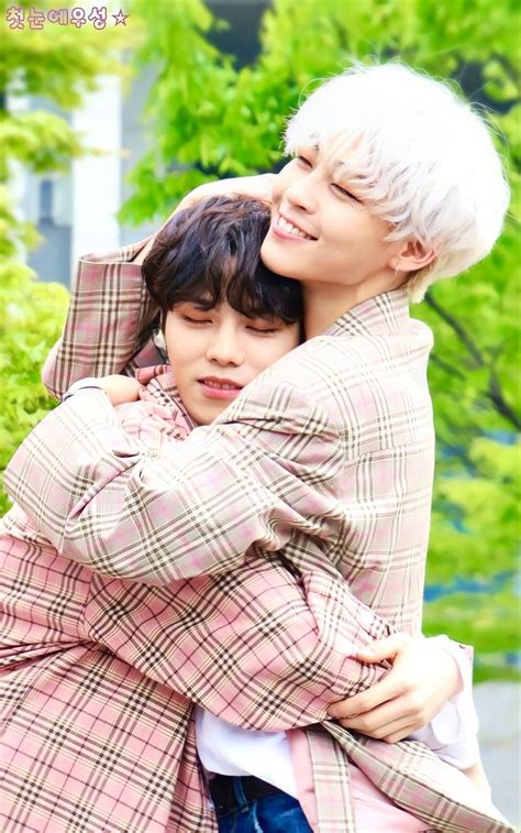 Two People Are Hugging Each Other In Front Of Some Trees And Bushes One Is Wearing A Plaid Coat