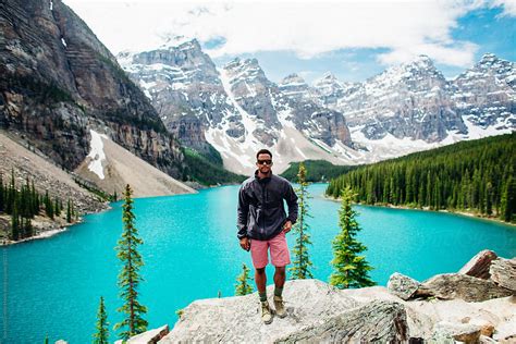 A Man Standing In Front Of A Blue Lake With A Mountain Landscape By