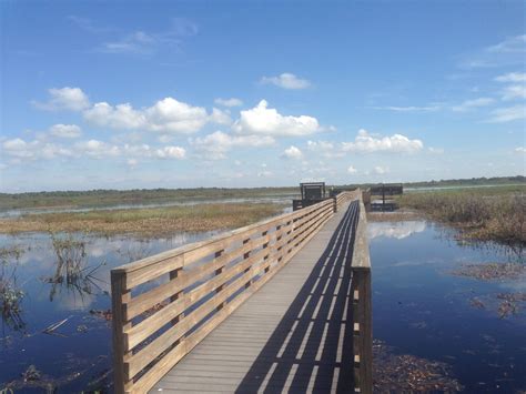 A Wooden Bridge Over Water With Benches On The Other Side And Clouds In
