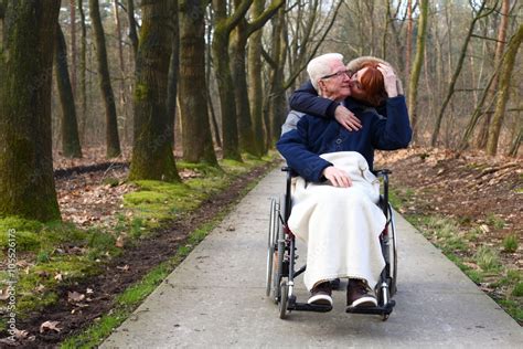 Kiss Young Woman Kissing An Old Man In A Wheelchair On His Cheek Stock