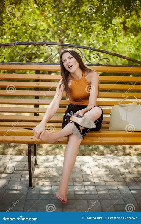 The Girl Tired Of Heels Takes Off Her Shoes From Her Feet And Sits Barefoot On A Park Bench