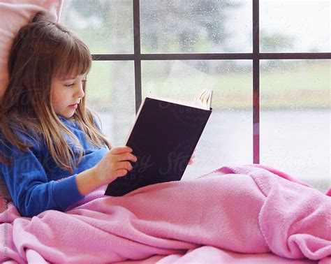 A Young Girl Sits In A Window Seat Reading A Book On A Rainy Day By