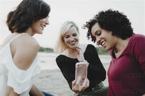 Cheerful Female Friends Video Conferencing While Sitting At Lakeshore