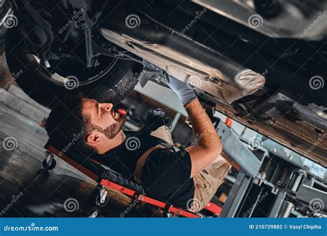Mechanic In Uniform Lying Down And Working Under Car At Auto Service