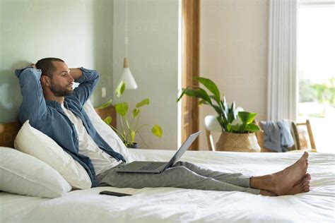 Thoughtful Man Sitting With Laptop On Bed At Home Stock Photo