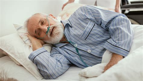 Sick Elderly Man Wearing Blue Pajama Lying In Bed At Nursing Hom